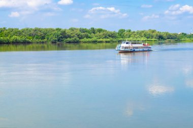 Beyaz bir turist gemisi, Omsk 'taki Irtysh Nehri boyunca kıyıdaki yeşil ağaçların arka planına doğru yavaşça ilerliyor. Bulutlar suyun sakin yüzeyine yansıyor. Yan görünüm - seyahat, turizm, su eğlencesi