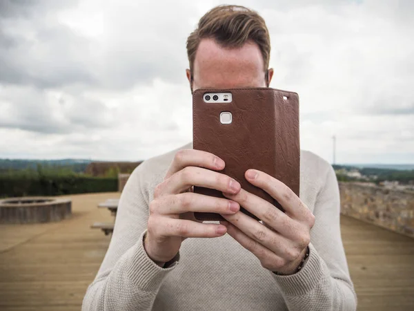 Young man with brown hair standing outdoor and taking a picture with his smartphone which has a brown leather cover