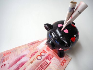 Black piggy bank standing next to Chinese 100 rmb banknotes on a white wooden background. The pig symbols fortune in the Chinese zodiac.