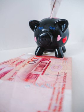 Black piggy bank standing next to Chinese 100 rmb banknotes on a white wooden background. The pig symbols fortune in the Chinese zodiac.