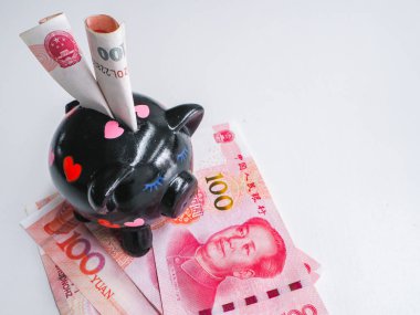 Black piggy bank standing next to Chinese 100 rmb banknotes on a white wooden background. The pig symbols fortune in the Chinese zodiac.