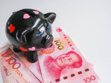 Black piggy bank standing next to Chinese 100 rmb banknotes on a white wooden background. The pig symbols fortune in the Chinese zodiac.