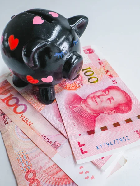 Black piggy bank standing next to Chinese 100 rmb banknotes on a white wooden background. The pig symbols fortune in the Chinese zodiac.