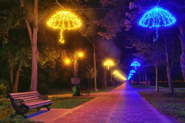 The alley in a night park with pavement, benches and flowerbeds is Illuminated with lanterns in the form of an umbrella. (city of Lutsk, Ukraine).