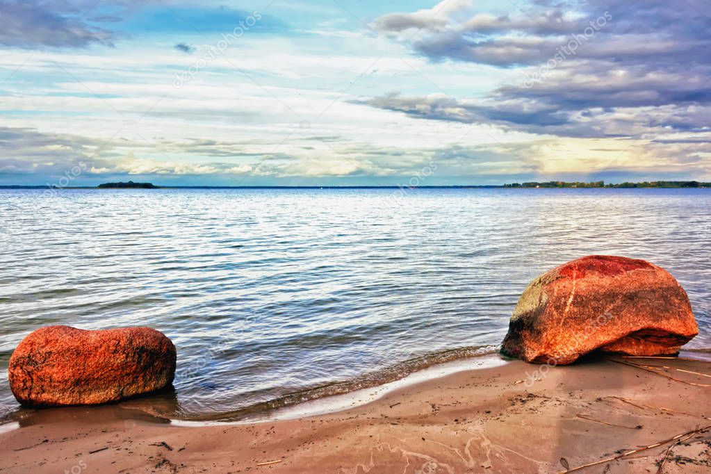 Sunset over the lake - a sandy beach with two stones, cloudy sky, the opposite shore, water with small waves.