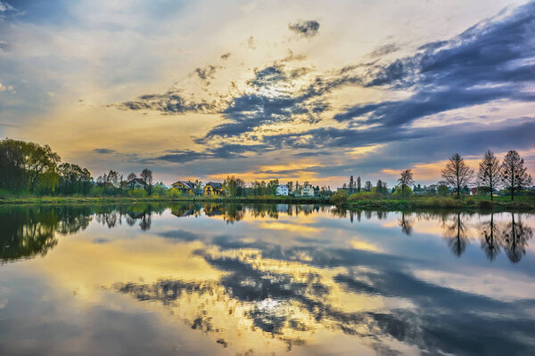 The sunset over the lake. Sky with clouds, trees and houses reflected in the water.