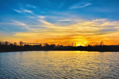 Göl kıyısında güzel bir gün batımı. Suya yansıyan ev ve ağaçların siluetleri.