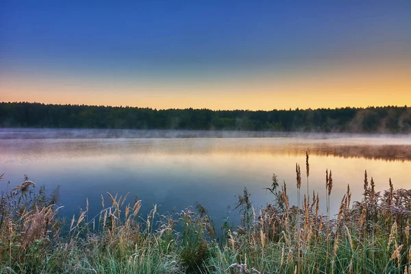 Autumn landscape - morning twilight over the lake. Fog over the water, the grass covered with frost.