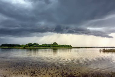 Gölün üzerinde fırtına bulutları ve çimen ve ağaçlarla kıyı şeridi.