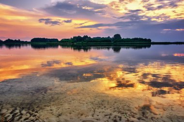 Gölün kıyı şeridinde gün batımı, bulutlar sakin suya yansıyor..