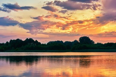Gölün kıyı şeridinde gün batımı, bulutlar sakin suya yansıyor..
