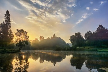 Yaz manzarası - ağaçlı ve çalılı küçük bir nehrin kıyısı sisli gündoğumunda.
