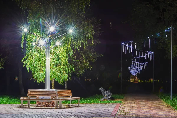 The alley in a night park with pavement, benches and flowerbeds is Illuminated with lanterns.