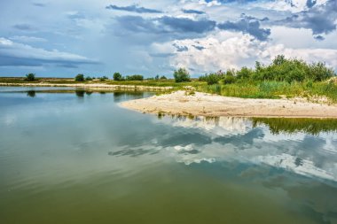 Fırtınada ağaçlar, çalılar ve çimenler olan nehir kıyısı. Fırtına bulutları suya yansıyor..