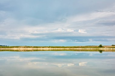 Bulutlu bir günde ağaçlarla, çalılarla ve otlarla dolu bir nehir kıyısı. Fırtına bulutları suya yansıyor..