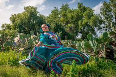 Renkli kurdeleli turkuaz bir elbiseyle Meksikalı bir halk dansçısının canlı portresi mavi gökyüzünün altında kaktüs ve ağaçlarla neşeyle dans ediyor..