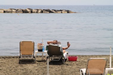 Güneşli bir günde kumlu bir plajda denizin önünde bir şezlong üzerinde yatan bir akıllı telefon ile tanınmayan kadın