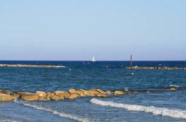 Taş kopuşları ile lacivert denizin panoramik manzarası, ufukta beyaz bir yelkenli tekne ve Limasol sahilinden açık mavi gökyüzü 