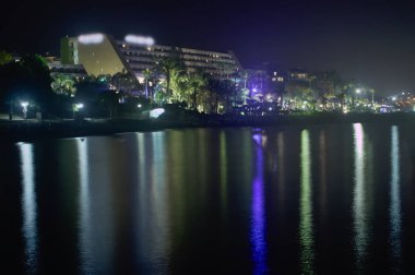Bir plaj otelinin yakınındaki ışıklı bir sahilin renkli gece ışıklarının yansıması ile deniz suyu