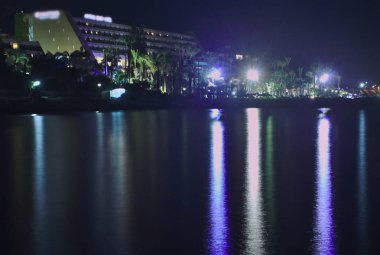 Bir plaj otelinin yakınındaki ışıklı bir sahilin renkli gece ışıklarının yansıması ile deniz suyu