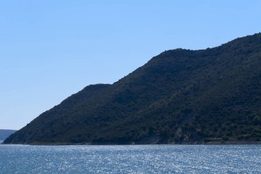 Green woody mountain slope with the clear blue sky above and the glittering lake waters below on a sunny day