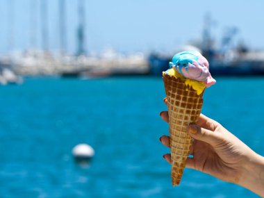 Bulanık mavi deniz ve teknelerin önünde renkli dondurmalı waffle külahı hazır.