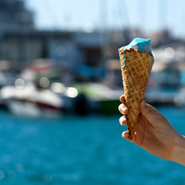 Bulanık mavi deniz ve teknelerin önünde dondurmalı waffle külahının yakın çekimi.