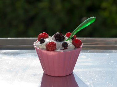 Taze böğürtlenli pembe bir kase yoğurt ve güneş ışığında parlayan plastik kaşık. 