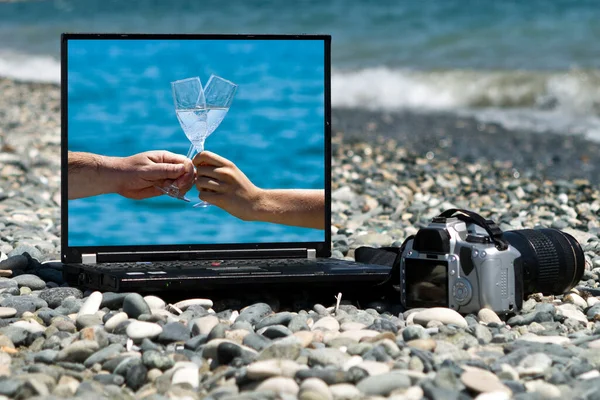 Laptop with picture of male and female hands holding two glasses of champagne on its monitor and camera laying on sea beach among gray stones