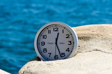 Yüzünde Carpe Diem yazan yuvarlak saat mavi denizin önündeki beyaz kayanın üzerinde yatıyor. 