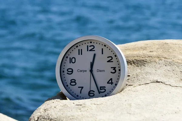 Yüzünde Carpe Diem yazan yuvarlak saat mavi denizin önündeki beyaz kayanın üzerinde yatıyor. 