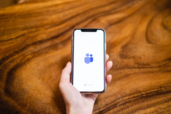 BANGKOK, THAILAND - 17 Mayıs 2020: Bir kullanıcı ahşap masadaki akıllı telefon ekranında Microsoft Ekip uygulamasını açar. Microsoft Takımları evden çalışmak için uygun popüler bir uygulamadır.