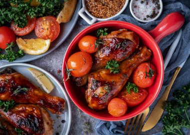 baked chicken legs in a barbecue sauce with tomatoes in a red frying pan, top view