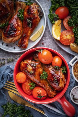 baked chicken legs in a barbecue sauce with tomatoes in a red frying pan, top view