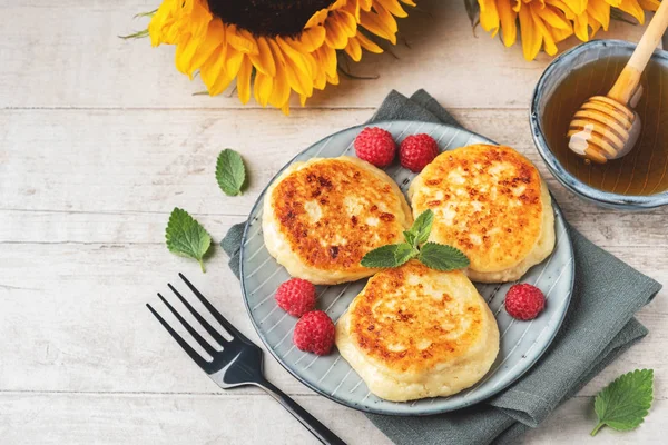 Cottage cheese pancakes with raspberries and mint leaves on a plate, wooden light background, top view, copy space. Food flat lay with sunflowers, pancakes and honey.
