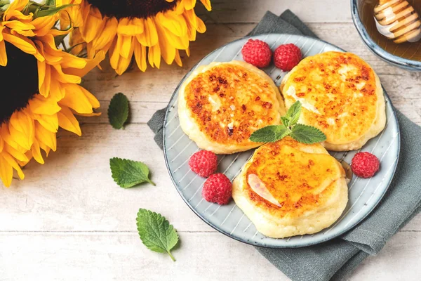 Cottage cheese pancakes with raspberries and mint leaves on a plate, wooden light background, copy space. Food flat lay with sunflowers, pancakes and honey.