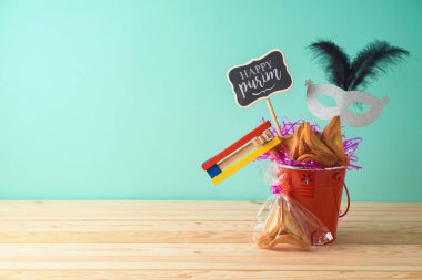 Jewish holiday Purim background with bucket, carnival mask, noisemaker and hamantaschen cookies on wooden table. Creative Purim basket idea clipart