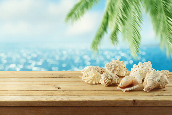 Summer background with wooden table, seashells nad palm tree over sea beach bokeh