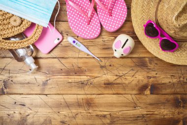 Yeni normal konsept. Seyahat bütçesi konsepti, domuz kumbarası, tıbbi maske, termometre, şapka ve terlikler ahşap masada. Yukarıdan üst görünüm