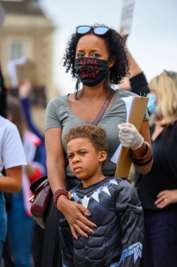 Richmond, Kuzey Yorkshire, İngiltere - 14 Haziran 2020: Güçlü bir kadın lider, Kuzey Yorkshire, Richmond 'da düzenlenen BLM protestosunda Siyah Yaşamlar Önemli PPE maskesi takıyor ve süper kahraman oğluna sarılıyor