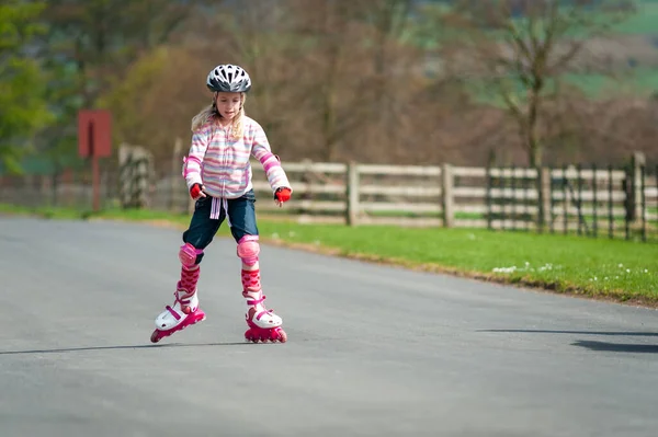 A young girl on roller blades and wearing protective equipment skates along a country road.