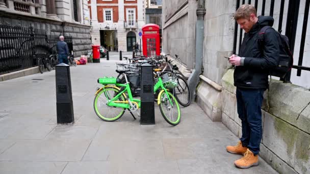 ロンドン 2020年2月4日 スマートフォンを操作し ライム電動アシスト自転車を見ている男 背景にはレッド ロンドン ダブル デッカー バスが通る — ストック動画