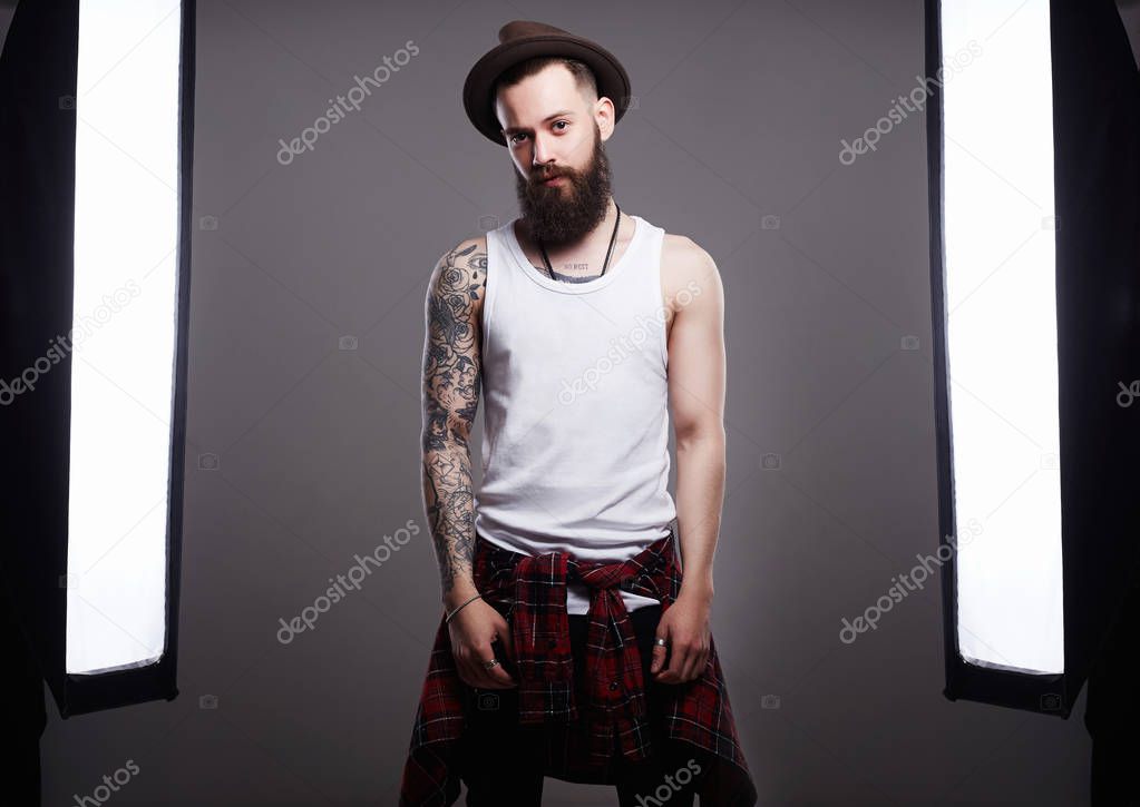 Handsome Bearded Man in Hat. Brutal hipster boy with tattoo posing in a photo studio in the middle of a photo of equipment