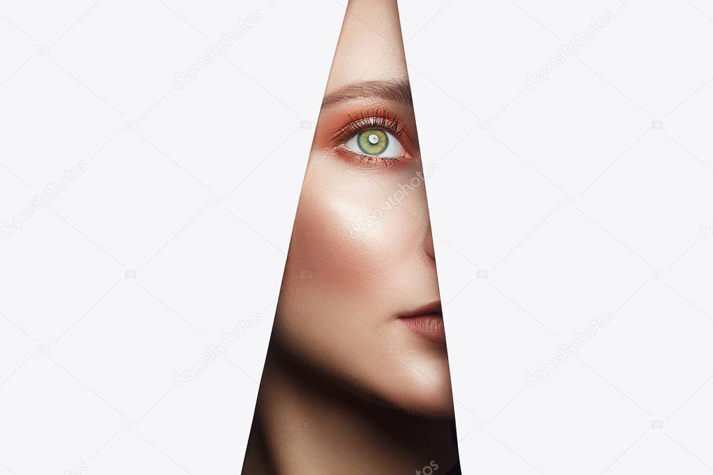 Face of a young beautiful woman with a bright makeup. make-up artist concept. A girl with beautiful bright green eyes with shining shadows, looks into the hole of white paper