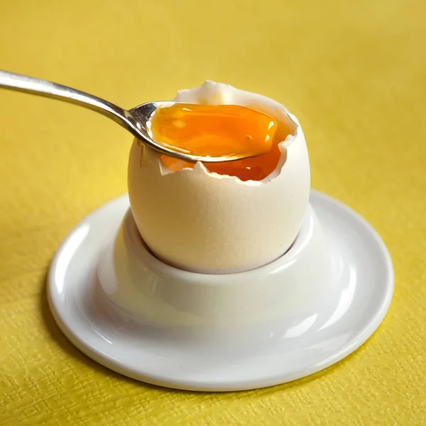 Soft-boiled egg in a porcelain egg cup with a little spoon on yellow background.