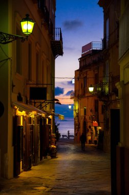 Night evening streets with bright lanterns lamps on buildings with balconies, silhouette of walking man and colorful sky with clouds on background, Tropea town, Vibo Valentia, Calabria, Southern Italy clipart
