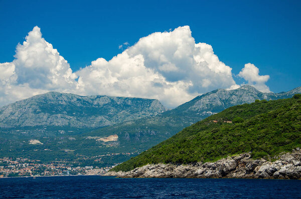 View of Boka Kotor bay water, town of Herceg Novi and Mount Orjen of Dinaric Alps mountain range in front of blue sky with white clouds, Montenegro