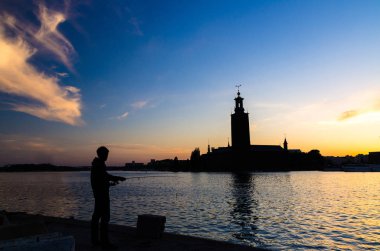 Olta kamışı ve Stockholm City Hall Kungsholmen Adası Belediye Meclisi Binası, günbatımı, alacakaranlık, alacakaranlık mavi sarı turuncu gökyüzü arka plan, İsveç ile Stadshuset ile balıkçı silüeti