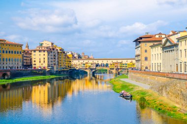 Floransa şehrinin tarihi merkezinde nehir kıyısı yakınında su ve tekneler yansıtan Arno Nehri mavi üzerinde renkli binalar evler ile Ponte Vecchio köprü, mavi gökyüzü beyaz bulutlar, Toskana, İtalya