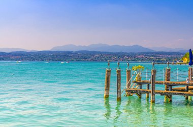 Mavi masmavi turkuaz suyu ve ahşap iskele rıhtımı olan Garda Gölü. Dağ sıraları, mavi gökyüzü arka planı, Sirmione kasabası, Lombardy, Kuzey İtalya.